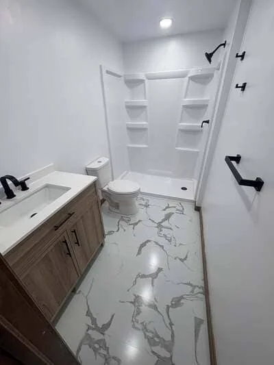 Bathroom with marble tile floor, white vanity, toilet, and tiled shower surround with shelves